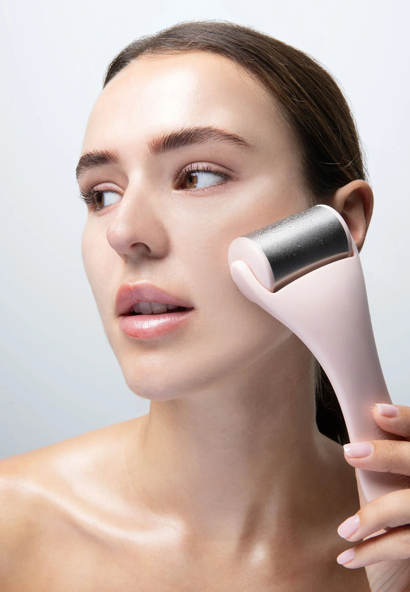 Pink facial ice roller with stainless steel head being used on woman’s cheek.