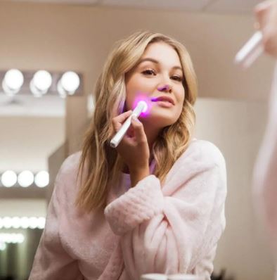 White handheld red and blue light therapy pen illuminating woman's cheek in bathroom mirror reflection.