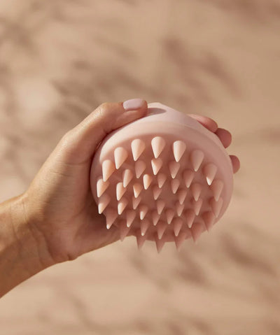 Hand holding a soft pink scalp massager with flexible conical silicone bristles against a beige background.
