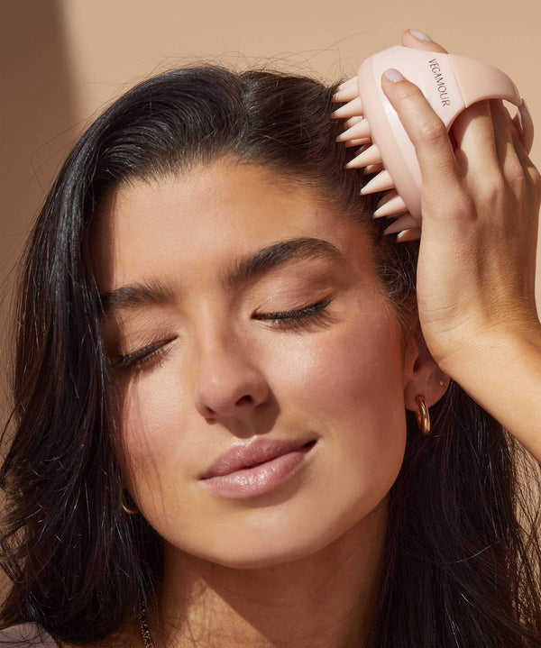 Woman using light pink scalp massager with soft silicone bristles on dark hair.