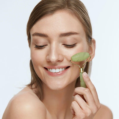 Woman using green jade facial roller on cheek with bare shoulders and smiling expression.